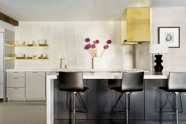 Contemporary open-plan kitchen with a long dark island and three black bar stools, pale flat-panel cabinetry, open timber-toned shelving, a light textured backsplash, and integrated appliances. Minimal decorative styling includes a sculptural black lamp and a simple floral arrangement.