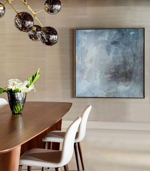 A refined dining area featuring a rounded dark timber dining table, sculptural white upholstered dining chairs, and a large abstract artwork on a pale textured wall. A polished brass and smoked-glass pendant light hangs overhead, while a dark vase with white flowers provides a natural accent.