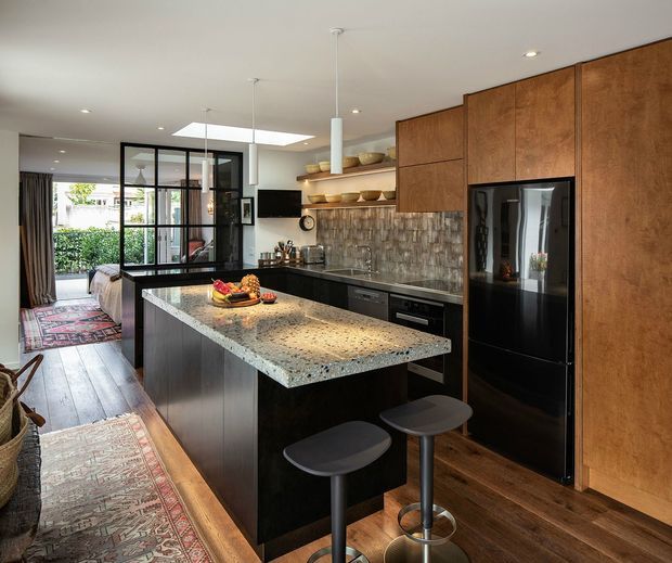 A contemporary open-plan kitchen with a large dark island, pale speckled stone benchtop, black appliances, and extensive warm timber cabinetry. Open shelving displays bowls above a textured tiled splashback. Two pendant lights hang over the island, with timber flooring and patterned rugs extending toward a glazed bedroom or living area beyond.