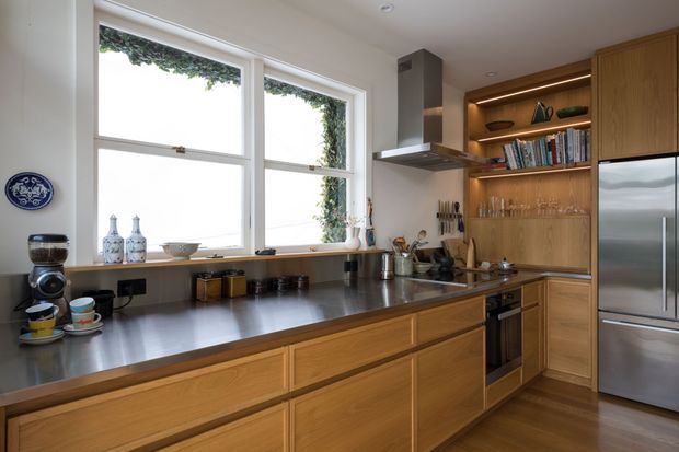 A warm, compact contemporary kitchen with extensive light timber cabinetry, dark stone-look benchtops, integrated open shelving, stainless-steel appliances, and a large double window above the work surfaces. Under-shelf lighting highlights books and glassware, while the island-style run provides generous preparation space.
