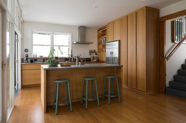 Warm contemporary kitchen with extensive timber cabinetry, a central island with three stools, stainless steel appliances, a large window over the rear benchtop, and an adjacent carpeted staircase. The room has timber flooring, white walls, and abundant natural light.