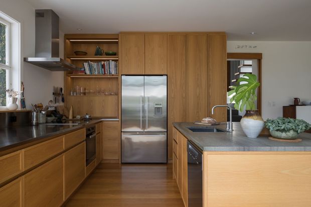 Warm contemporary kitchen with extensive natural timber cabinetry, dark stone benchtops, stainless-steel appliances, open shelving, and a central island with integrated sink. A large window and glazed doorway bring in natural light, while indoor plants and timber flooring add an organic feel.