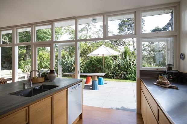 Bright, open-plan kitchen viewed toward a paved outdoor terrace and lush garden through extensive white-framed glazing. The kitchen features warm timber cabinetry, dark stone benchtops, a deep undermount sink, integrated dishwasher, and a secondary work surface with small appliances. A dining or living area is partially visible at left, while the outdoor area includes a table, stools, and umbrella.