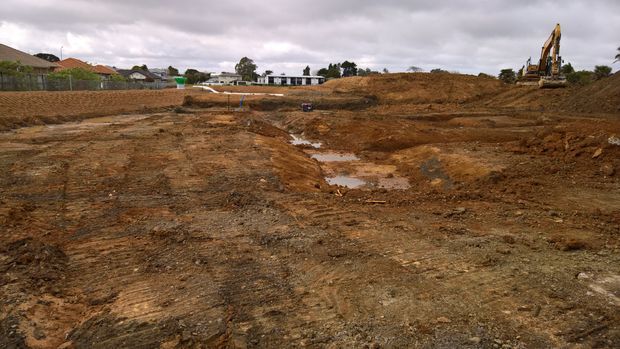 A wide outdoor view of a cleared and excavated construction site, with exposed brown soil, muddy puddles, earthmoving tracks, and an excavator on the right. Residential properties are visible along the distant edge under an overcast sky.
