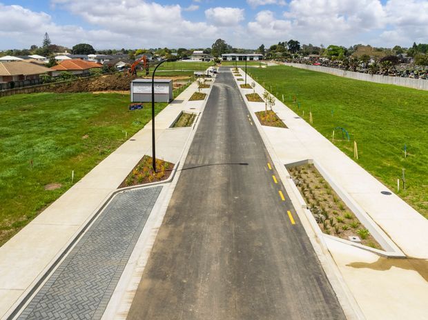 Aerial view of a newly developed suburban street with fresh asphalt paving, concrete footpaths, landscaped planting beds, street lighting, and broad grassed areas. Low-rise residential properties and construction activity are visible around the site under a partly cloudy sky.