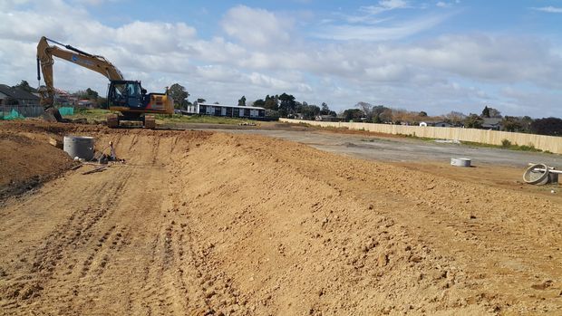A broad outdoor construction site with freshly graded sandy soil, an excavator, exposed drainage infrastructure, and a paved area beyond. Low buildings and fencing sit in the background under a partly cloudy sky.