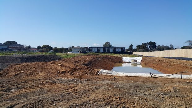 A wide outdoor view of a residential construction site with freshly excavated earth, a temporary plastic-lined water or drainage basin, construction fencing, and modern homes in the background under a clear blue sky.