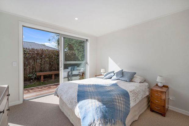 Bright, neutral bedroom with a double bed dressed in white bedding and a blue throw, pale walls, beige carpet, and a timber bedside table. Large glazed sliding doors open to a small timber deck and planted outdoor area, bringing in abundant natural light.