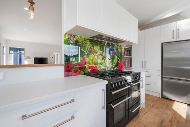Bright contemporary kitchen with white flat-panel cabinetry, a white benchtop, stainless-steel refrigerator, and a large black freestanding range. A vivid tropical botanical glass splashback forms the main feature, while timber flooring and a timber-trimmed pass-through connect the kitchen to the living area beyond.