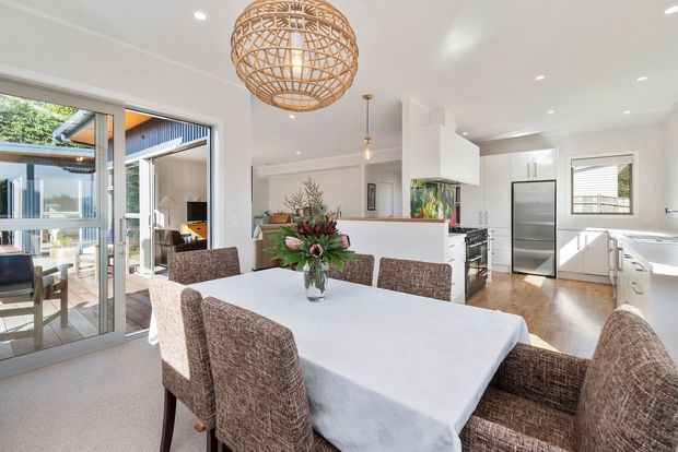 Bright open-plan dining and kitchen interior with a large rectangular dining table, woven upholstered dining chairs, white cabinetry, stainless-steel refrigerator, timber flooring, and a prominent woven pendant light. Sliding glass doors open to an outdoor area, bringing in abundant natural light.