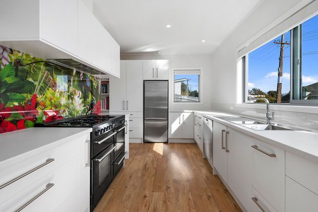 Bright contemporary galley kitchen with extensive white cabinetry, a stainless-steel range and refrigerator, a large window above the sink, warm timber flooring, and a colourful botanical splashback behind the cooktop.