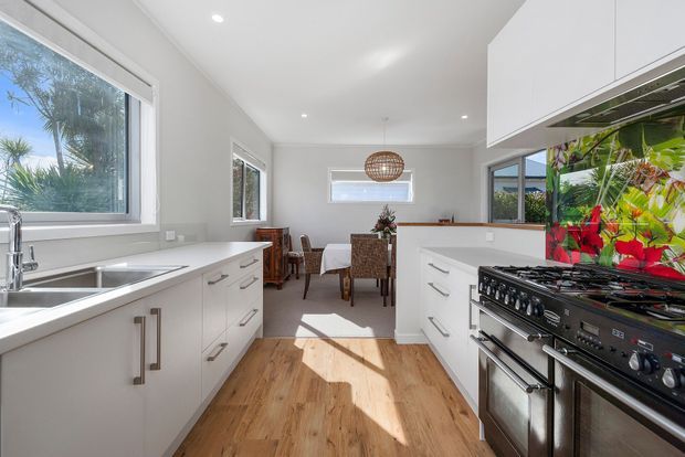 Bright contemporary kitchen with white cabinetry, stainless-steel fixtures and appliances, timber-look flooring, and a central dining area. Large windows provide abundant natural light, while a colourful botanical splashback adds a strong feature element behind the freestanding range cooker.