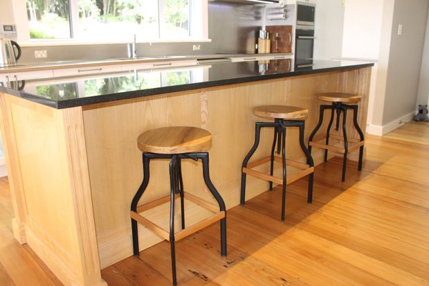 Bright contemporary kitchen featuring a long timber-clad island with a dark stone benchtop, integrated seating for three, polished timber flooring, stainless steel appliances, and large windows.