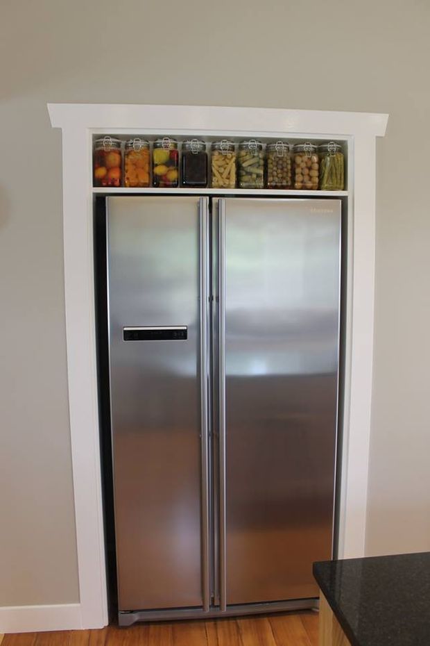 A stainless-steel side-by-side refrigerator is recessed into a white-trimmed kitchen wall, with an open display shelf above holding jars of preserved food. The lower right corner shows a dark stone kitchen benchtop, and timber flooring is visible below.