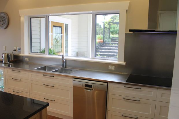A bright, practical kitchen with white shaker-style cabinetry, a dark polished island benchtop, stainless-steel work surfaces and appliances, and a wide window overlooking outdoor steps and greenery. The room has a clean traditional-to-transitional character with abundant natural light.