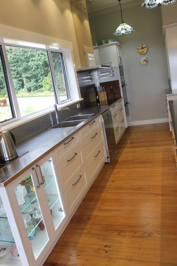 A narrow, traditionally styled kitchen viewed along a long run of cream shaker-style cabinetry. The room features a stainless-steel benchtop and double sink beneath a large window, integrated appliances, dark timber splashback accents, warm timber flooring, glass-front display cabinetry, and decorative pendant lights. The space is bright and functional, with greenery visible outside the window.