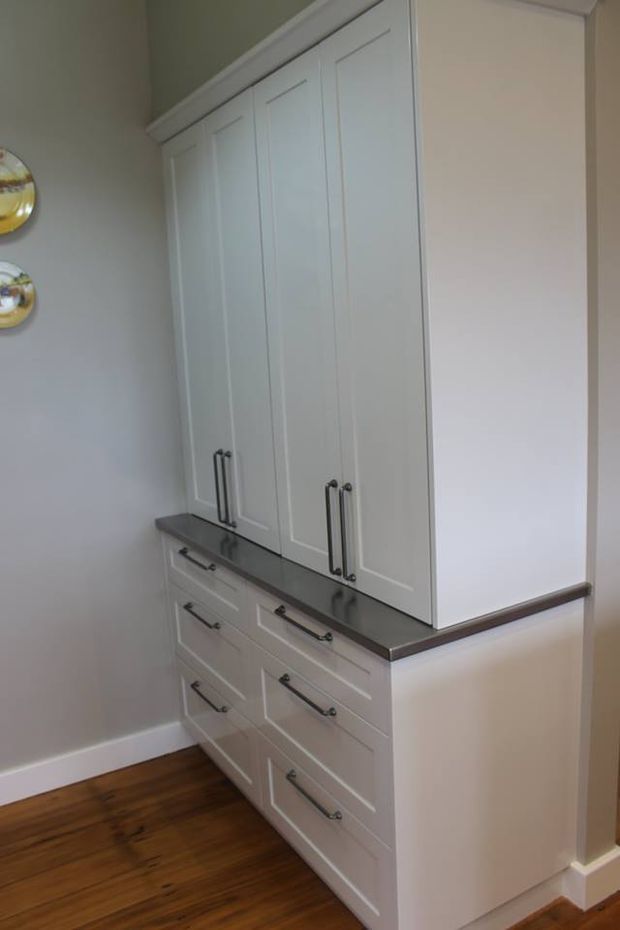 A compact built-in white storage unit with Shaker-style paneled cabinet doors and multiple drawers, topped by a dark stone or engineered-stone counter. Traditional metal pull handles, white walls and trim, and warm timber flooring create a clean, understated built-in joinery installation.