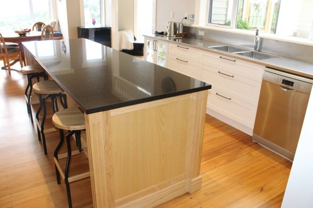 Bright, spacious kitchen with a large dark stone island and timber-panelled base, integrated seating, white shaker-style cabinetry, stainless-steel sink and dishwasher, and warm timber flooring. A dining area is visible beyond the kitchen.