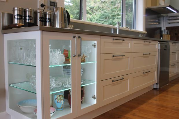 A bright, traditional-style kitchen with a long built-in island or cabinet run. The cabinetry is painted warm beige with framed drawer fronts, dark metal pulls, and illuminated glass-front display cupboards holding glassware and ceramics. A dark benchtop, stainless-steel appliances, timber flooring, and a large window overlooking greenery complete the space.
