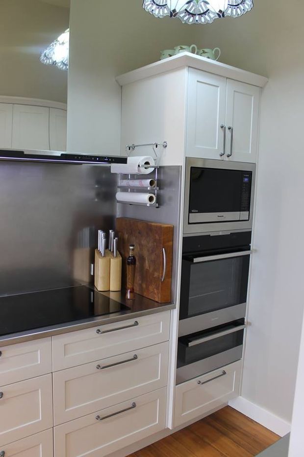 A compact, well-organised kitchen featuring white Shaker-style cabinetry, a black glass induction cooktop, integrated stainless-steel oven and microwave stack, dark benchtops, a reflective metallic splashback, timber flooring, and traditional decorative lighting. Open shelving and a wall-mounted paper-towel holder sit beside a timber chopping board and knife blocks.