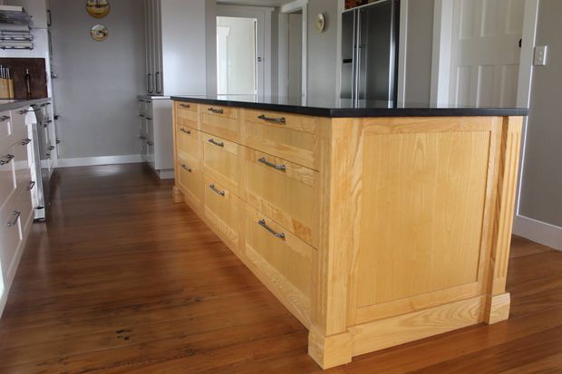 A traditional-style kitchen centred on a large custom timber island with paneled ends, framed drawers, and dark metal pulls. The room has warm timber flooring, white cabinetry, grey walls, stainless-steel appliances, and a doorway leading to adjoining spaces.