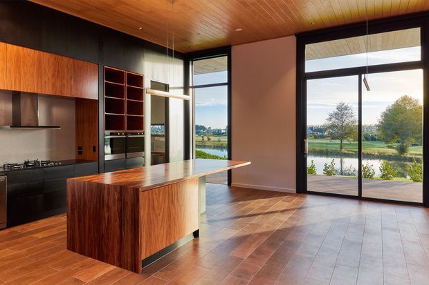 A spacious modern kitchen and open-plan living area with a large timber island, dark built-in cabinetry, and full-height black-framed glazing. The room features a warm timber-lined ceiling and timber-look tiled flooring, with views over a landscaped waterfront or lakeside setting. Strong natural light fills the space through the expansive glass doors and windows.