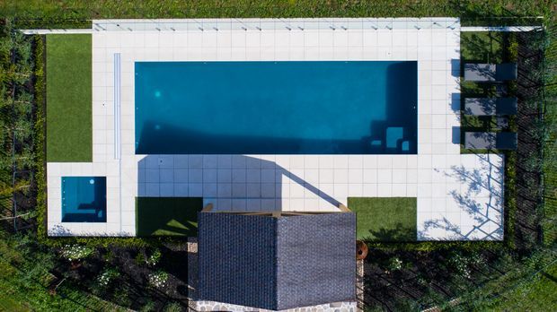 Aerial top-down view of a landscaped residential property centred on a large rectangular swimming pool, with a smaller adjacent plunge pool, pale tiled surrounds, geometric lawn areas, a tiled outdoor terrace with seating, and a dark pitched-roof building at the lower edge.