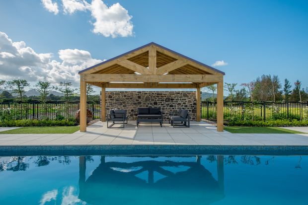 A freestanding timber-framed pool pavilion with an open gable roof, exposed rafters and beams, and a rustic stone feature wall. Outdoor lounge furniture sits on a tiled terrace beside a swimming pool, with landscaped grounds and fencing beyond.