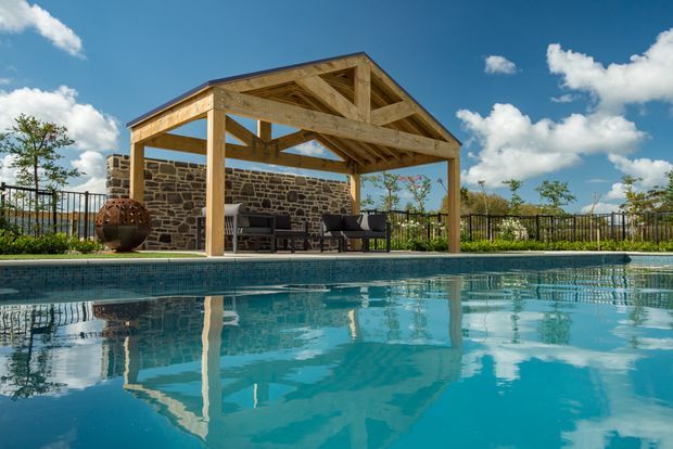 An outdoor swimming pool occupies the foreground, reflecting a timber-framed, gable-roofed pool pavilion. The pavilion contains outdoor lounge seating and is backed by a rustic stone wall and metal fencing, with landscaped surroundings and a bright blue sky.