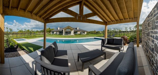 A covered outdoor entertaining area with a timber-framed, pitched roof and exposed rafters, furnished with dark outdoor sofas and a coffee table. It overlooks a rectangular swimming pool, paved terrace, landscaped lawn, and a single-storey house, with a rough stone wall along the right side.