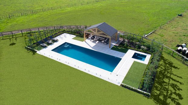 Aerial view of a landscaped outdoor pool area set within a large rural pasture. The rectangular swimming pool has a white paved surround and an attached shallow spa or plunge pool, with a timber-framed, pitched-roof pavilion containing outdoor seating at one end. Fencing and trellised planting define the perimeter, with livestock visible in the surrounding field.