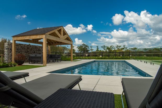 A landscaped outdoor pool area viewed from lounge chairs, featuring a rectangular swimming pool, broad pale paving, dark outdoor loungers, and a timber-framed pitched-roof pavilion with an outdoor seating area and stone wall backdrop. The setting appears spacious, sunny, and recreational.