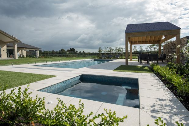 A landscaped outdoor pool area with two connected rectangular swimming pools, pale tiled paving, manicured lawn, and a timber-framed covered pavilion with outdoor seating. The scene has a spacious suburban or rural setting under a dramatic cloudy sky.