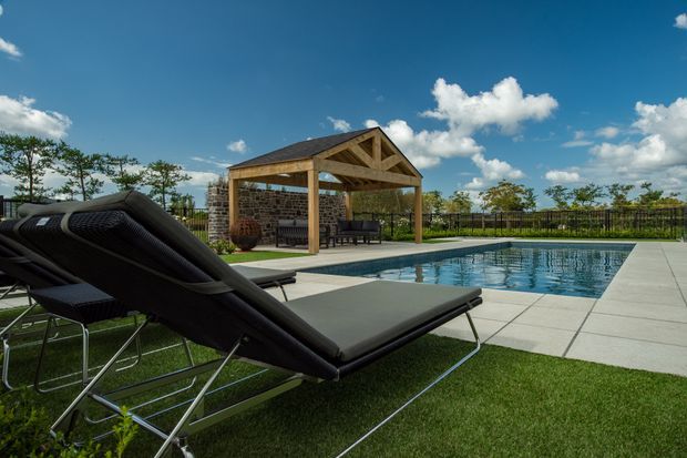 A landscaped outdoor pool area with a rectangular swimming pool, pale paved surround, black reclining sun loungers, and a timber-framed, gabled pavilion with stone-clad walls and outdoor seating. The scene is bright and open, with a lawn, fencing, and blue sky in the background.