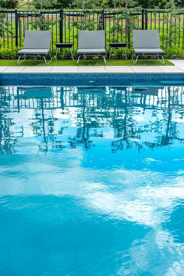 A bright outdoor swimming pool with clear turquoise water in the foreground, bordered by a narrow pale tiled coping and a strip of artificial or manicured grass. Three grey lounge chairs and small black side tables sit behind a black metal fence, with lush greenery beyond. The image focuses on the pool and relaxed poolside setting.