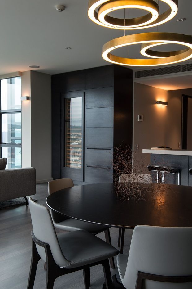 A refined contemporary open-plan dining and living area with a large round black dining table and upholstered chairs in the foreground. Dark built-in wall joinery conceals storage and a door, while a compact bar or kitchen zone sits behind it. Two large circular pendant lights with warm illuminated rings create a strong focal point. Floor-to-ceiling glazing brings in daylight and urban views, and the palette is dominated by charcoal, black, grey, white, and warm metallic tones.