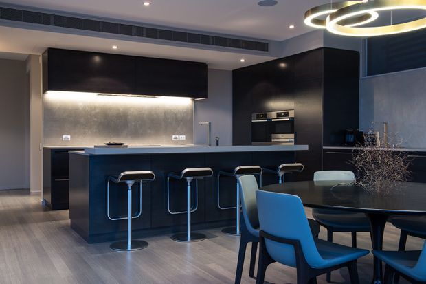 A sophisticated open-plan kitchen and dining area with dark glossy cabinetry, a long island with integrated seating, a metallic textured splashback, and a contemporary dining table with blue upholstered chairs. Recessed ceiling lights and a sculptural circular pendant create a refined, modern atmosphere.