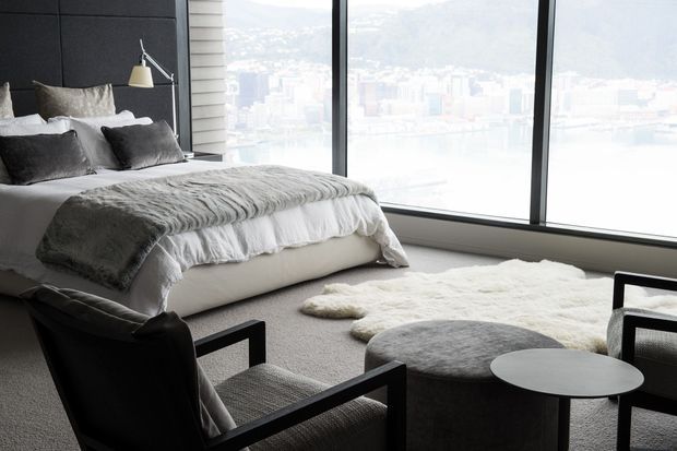 A sophisticated bedroom with a large upholstered bed, dark padded feature wall, layered grey bedding, black-framed floor-to-ceiling glazing, and a soft white rug. A pair of dark lounge chairs and round side tables occupy the foreground, while a bright urban and mountainous view is visible beyond the windows.