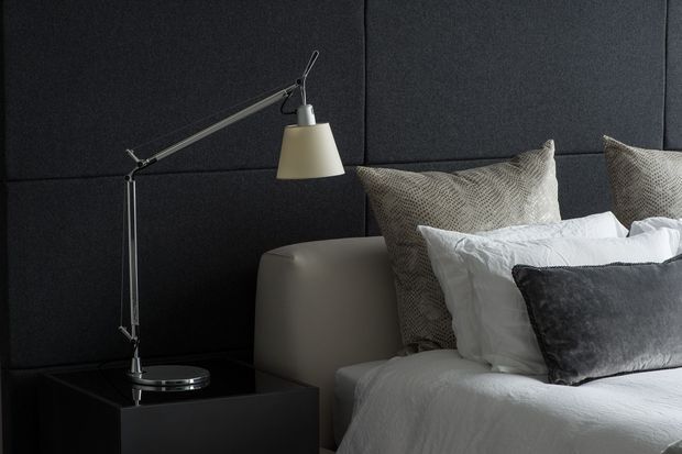 A moody, refined bedroom vignette featuring a bed with layered white and grey bedding, textured neutral pillows, a dark charcoal upholstered feature wall, and a minimalist articulated desk lamp on a dark bedside table. The room has a contemporary, softly luxurious feel with strong tonal contrast.