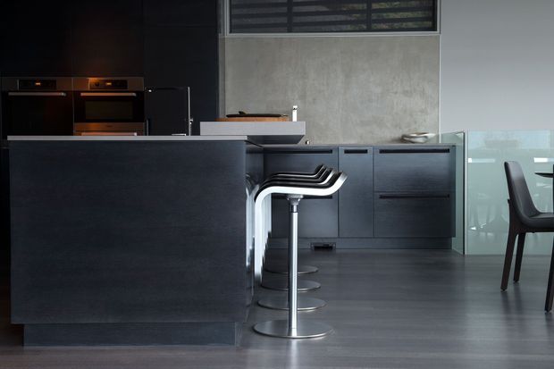 A sleek contemporary kitchen with dark matte cabinetry, a large island with a pale stone benchtop, integrated dark appliances, and sculptural chrome bar stools. The kitchen opens toward a dining area with dark seating and translucent glass screening. The space has a restrained monochromatic palette and polished, modern detailing.