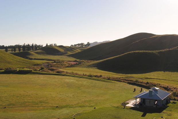 Aerial view of a small rural house set within expansive rolling green farmland, with sculpted grassy hills, sparse trees, fences, and open pasture surrounding the property. The home has a compact gabled roof and a simple detached form. Warm low-angle sunlight creates long shadows across the landscape.