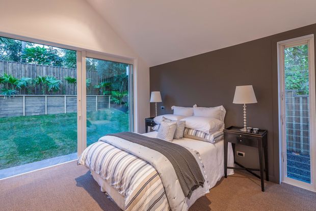 A bright, contemporary bedroom with a pitched ceiling, full-height glazing and sliding doors opening toward a private garden. The room features a neatly made bed with layered white and striped bedding, dark brown feature walls, beige carpet, and matching bedside tables with lamps.