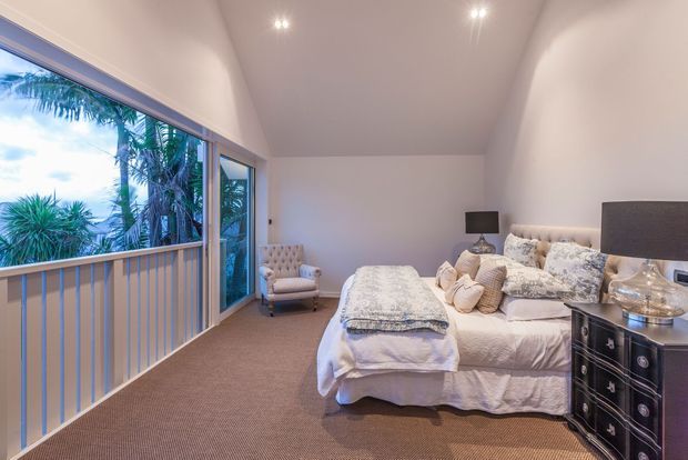A bright, spacious bedroom with a large bed dressed in white and patterned bedding, upholstered bedside furniture, bedside lamps, a dark chest of drawers, and warm beige carpet. Floor-to-ceiling glazing and a balustraded opening overlook lush tropical vegetation, creating an airy indoor-outdoor feel.