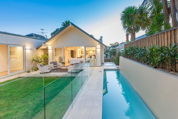 A landscaped residential courtyard viewed from outdoors, featuring a narrow rectangular swimming pool beside a pale paved path, frameless glass pool fencing, a manicured lawn, tropical planting and timber privacy fencing. The background shows a single-storey light-coloured house with a prominent gabled roof, glazed living area and warm interior lighting.