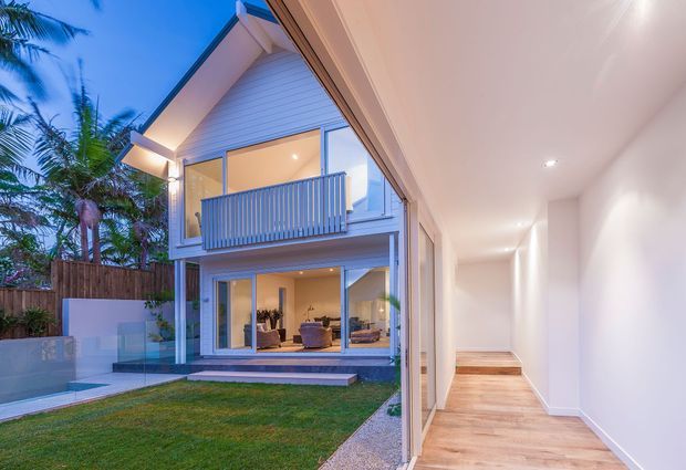 Evening view of a contemporary two-storey home with a light weatherboard facade, pitched gable roof, glazed lower-level living area, and a small upper balcony. The image is taken from a bright minimalist interior hallway opening toward a landscaped courtyard and the house exterior, with warm timber flooring and recessed lighting inside.