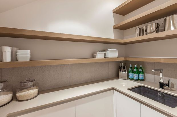 Compact contemporary kitchen with white handleless cabinetry, a pale engineered-stone benchtop, integrated dark sink, warm timber open shelving, and a neutral stone-look splashback. Open shelves display crockery, cookware, pantry jars, and bottled drinks.