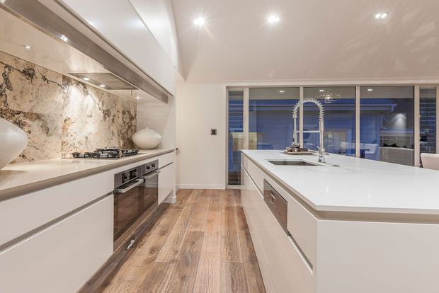 Bright contemporary kitchen with a large white island, integrated cabinetry, timber-look flooring, marble-pattern splashback, stainless-steel appliances, and expansive glazing overlooking an outdoor area.