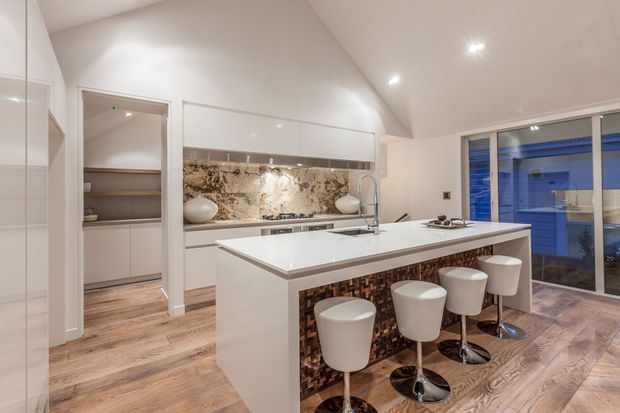 Contemporary open-plan kitchen with a large white island and integrated sink, seating for four on white bar stools, glossy white cabinetry, and a dramatic natural stone backsplash. Warm timber-look flooring and a textured mosaic feature panel add contrast, while recessed lighting and large sliding glass doors create a bright, polished space.