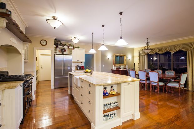 A spacious traditional kitchen and dining area with a large cream island, marble-look benchtop, white cabinetry, stainless-steel refrigerator, timber flooring, ornate cornices, pendant lights, and detailed draped window treatments. A formal dining table and chairs are visible beyond the kitchen.
