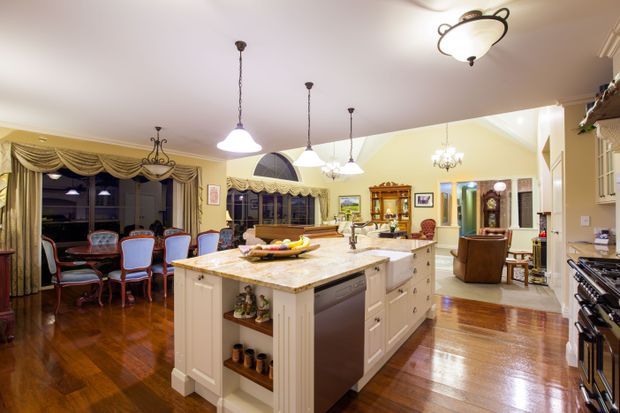 Spacious traditional open-plan kitchen and dining area with a large central island, white paneled cabinetry, a light stone benchtop, pendant lighting, polished timber floors, and ornate draped window treatments. The adjoining dining and living areas are visible, with classic furniture and decorative joinery.